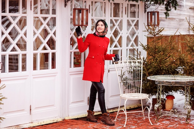 Modieuze vrouw in rode jas brengt in de winter tijd buiten door naast de witte veranda.