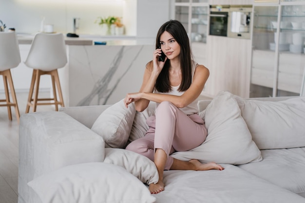 Moe Aziatisch meisje in roze broek en wit t-shirt zittend op de bank pratend via de telefoon opzij kijkend tegen wazige woonkamer Schitterend model praat met vriend verveeld door quarantaine Mooie mensen