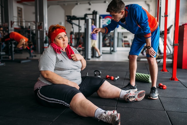 Moe dikke vrouw zit op de vloer, training met instructeur in de sportschool. Calorieën branden, zwaarlijvige vrouwelijke persoon in sportclub