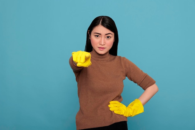 Moe gefrustreerde en ongelukkige aziatische huisvrouw die met de vinger naar voren wijst en gele handschoenen draagt, huisconcept schoonmaken, verdrietig gestresst dienstmeisje met negatieve gemoedstoestand