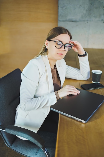 Moe jonge zakenvrouw zittend op kantoor aan een houten tafel achter een laptop Een jonge manager met een bril is het werk beu