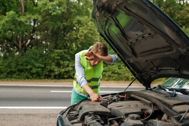 Moe jongeman bij de geopende motorkap fix probleem met motor, autopech. Kapotte auto of reparatie van voertuig, problemen met auto op snelweg