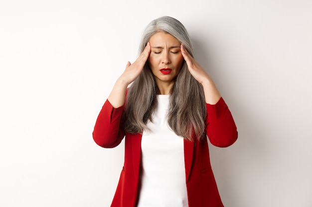 Moe senior zakenvrouw in rode blazer met hoofdpijn, hoofd aanraken en ziek voelen, staande met migraine tegen witte achtergrond.