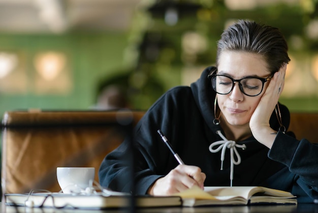 Moe snelkoppeling student in stijlvolle optische bril zittend aan houten tafel in cafetaria met kopje koffie en iets schrijven op de notebook