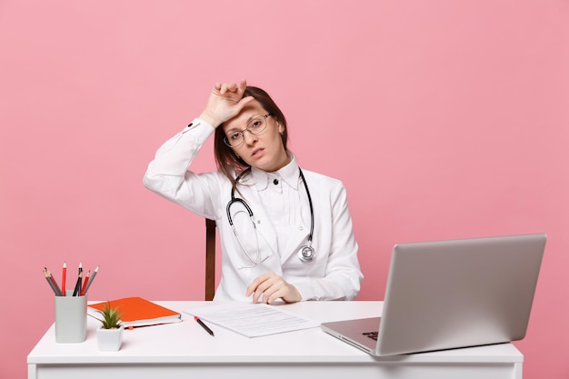 Moe trieste vrouwelijke arts zit aan bureau werk op computer met medisch document in ziekenhuis geïsoleerd op pastel roze muur achtergrond. Vrouw in medische toga glazen stethoscoop. Gezondheidszorg geneeskunde concept.
