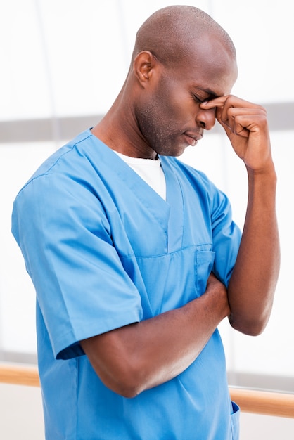 Moe voelen na een lange operatie. Portret van een depressieve jonge Afrikaanse arts in blauw uniform die zijn neus met de hand aanraakt en de ogen gesloten houdt