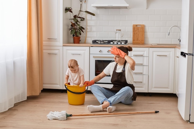 Moe vrouw poseren met baby baby tijdens het schoonmaken in een huis keuken problemen met huishoudelijke taken na de geboorte van een kind vrouw in schort zittend op de vloer en kijken naar kind in gele emmer