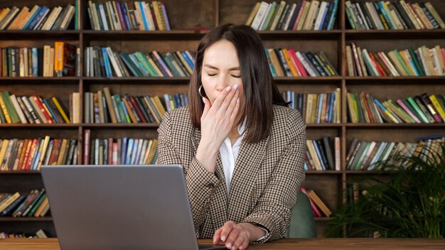 Moe zakenvrouw in bruine jas typen op grijze laptop en gegaap zittend op stoel aan tafel tegen houten rekken met boeken thuis