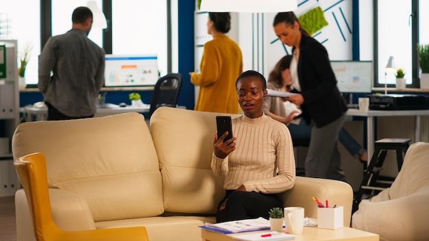 Moe zwarte vrouw praten met collega met behulp van video-oproep met smartphone met draadloze headset zittend op de bank in kantoor. Divers team dat nieuw financieel project plant in modern bedrijf