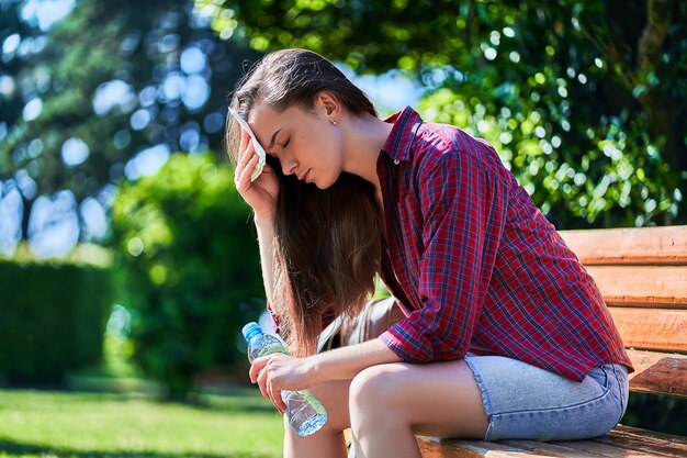 Moe zwetende vrouw met fles water rustend op een bankje en veegt haar voorhoofd met een servet in een park in warm zomerweer