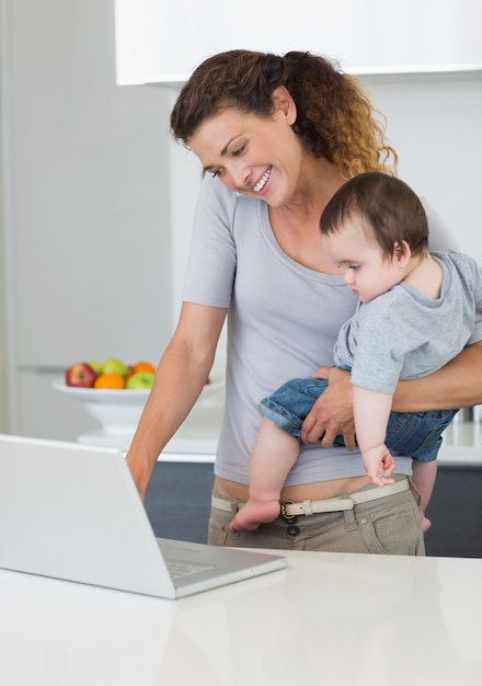 Moeder die laptop met behulp van terwijl het dragen van baby in keuken