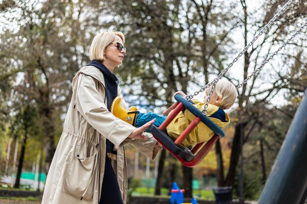 Moeder duwt haar baby jongen kind met gele regen laarzen en cape op schommel op de speeltuin