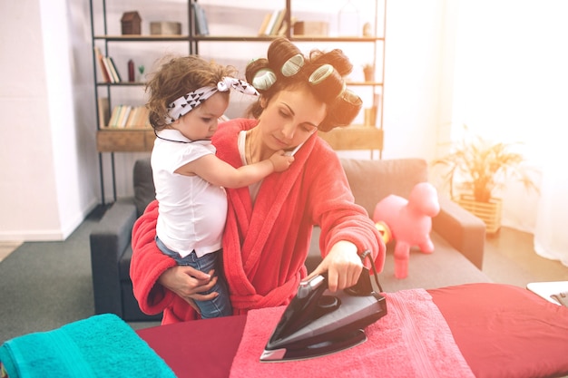 Moeder en baby samen bezig met huishoudelijk werk Strijken van kleding. Huisvrouw en kind huiswerk. Vrouw met klein kind in de woonkamer. Huisvrouw doet veel taken terwijl ze voor haar baby zorgt.