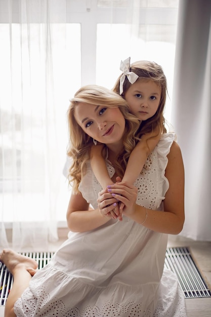 Moeder en dochter baby in witte jurken zittend op de vloer van het huis tegen de achtergrond van het raam