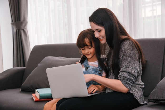 Moeder en dochter brengen samen tijd door in de woonkamer Moeder leert haar dochter online thuisonderwijs via e-learning op afstand voor studie op laptop