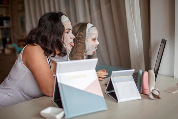 Foto moeder en dochter die thuis een gezichtsmasker aanbrengen met behulp van een spiegel en een laptop