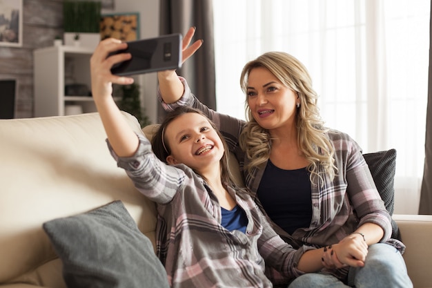Moeder en dochter in bijpassende kleding zitten op de bank en nemen een grappige selfie in de woonkamer.