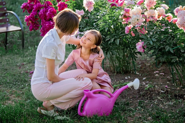 Moeder en dochter in een linnen roze jurk zorgen voor pioenrozen in de tuin, geven de bloemen water. Hij lacht perfect en lacht. Verlicht door de stralen van de avond ondergaande zon.