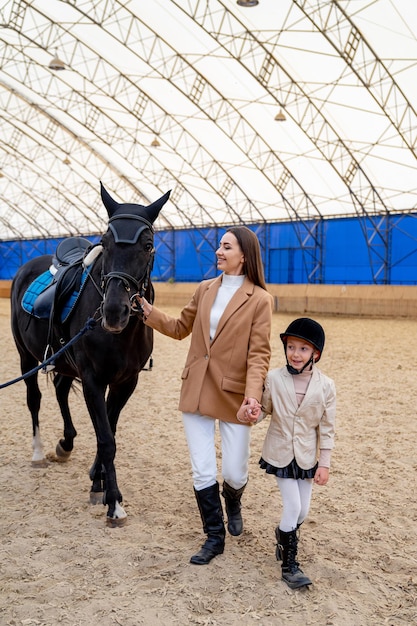 Moeder en dochter in helder pak en hoed lopen met een zwart paard in de overdekte arena Het concept van paardensport paardenfokken