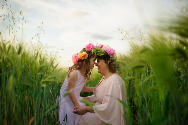 Moeder en dochter in roze jurken in een groen tarweveld. Op hun hoofd hebben ze bloemenkransen.