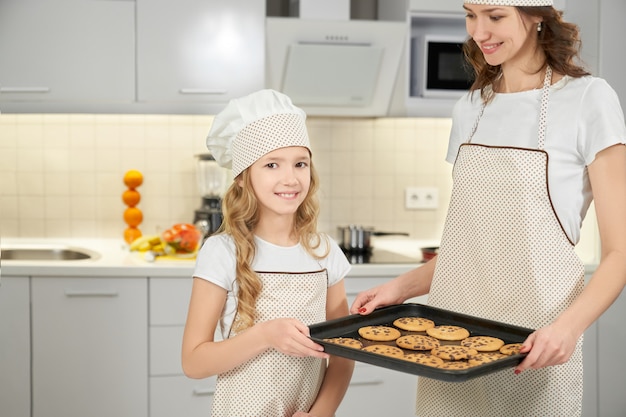 Moeder en dochter in schorten en chef-kok hoeden poseren met koekjes