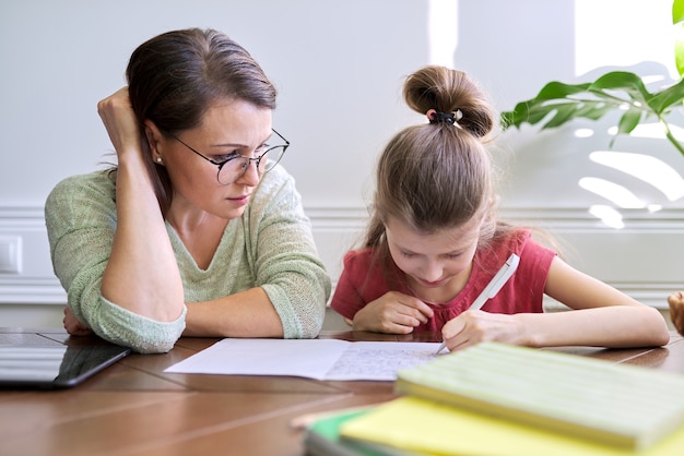 Moeder en dochter kind studeren samen thuis, zittend aan tafel, meisje schrijft in notitieblok. afstandsonderwijs, ouder helpt kind basisschoolleerling