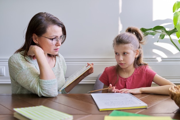 Moeder en dochter kind studeren samen thuis, zittend aan tafel. Vrouw met boek, meisje schrijft in notitieblok. Afstandsonderwijs, ouder helpt kind basisschoolleerling