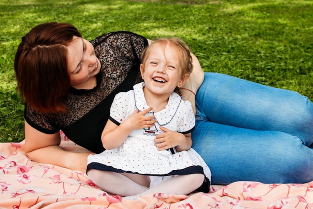 Moeder en dochter lachen en knuffelen zittend op een deken in het park op een open plek