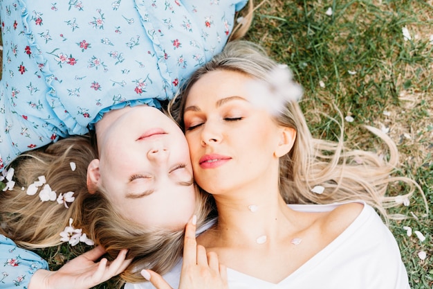 Foto moeder en dochter liggen op het gras en vallende boombloemen, blond haar, park of bos