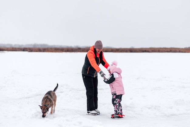 Moeder en dochter op winterwandeling Jonge vrouw en klein meisje lopen langs bevroren rivieroever hond speelt naast hen winterlandschap