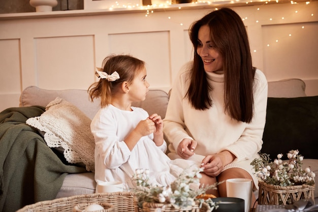 Moeder en dochter praten op de bank in de woonkamer ingericht voor Kerstmis. Jonge gelukkige familie viert Kerstmis. Gezellige Kerst