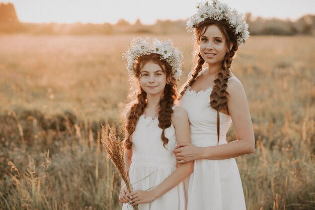 Moeder en dochter samen in witte jurken met vlechten en bloemenkransen in bohostijl op het gebied van de zomer bij zonsondergang