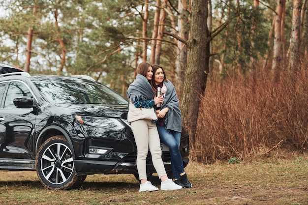 Moeder en dochter staan samen in de buurt van moderne zwarte auto buiten in het bos