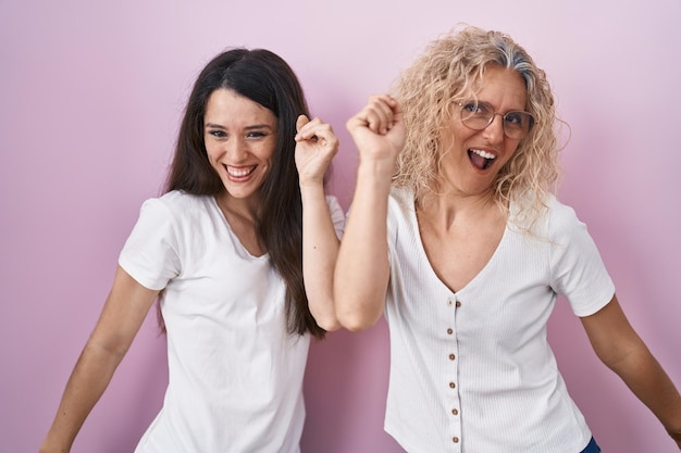 Moeder en dochter staan samen over een roze achtergrond, dansen blij en vrolijk, glimlachend bewegend, casual en zelfverzekerd luisterend naar muziek