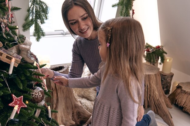 Moeder en dochter versieren de kerstboom. Portret liefdevolle familie in mooie vakantie DIY handgemaakte woondecoratie. Gezellige kamer versierd met kaarsen en kerstboom met cadeautjes eronder