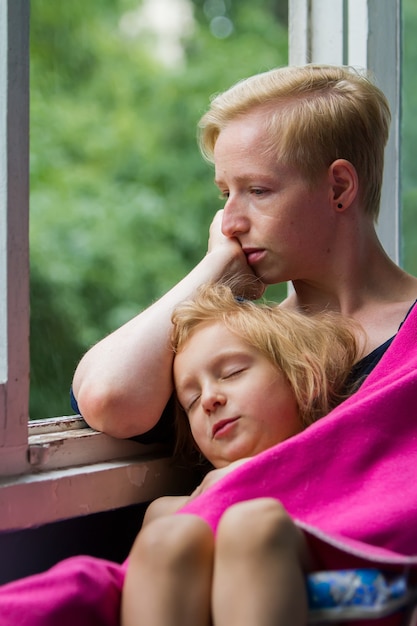 Foto moeder en dochter zitten bij het open raam, de moeder houdt haar slapende dochter in een deken over zich heen