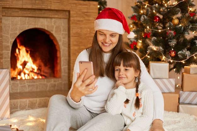 Moeder en dochter zitten in de buurt van de kerstboom, knuffelen, communiceren via videogesprek, plezier maken.