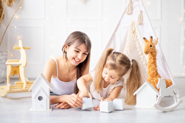 Moeder en dochtertje spelen thuis in een lichte kinderkamer tussen houten speelgoed glimlachend en plezier gelukkig moederschap