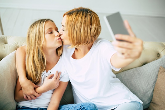 Moeder en haar dochtertje knuffelen, poseren en maken selfie foto op smartphone terwijl ze thuis op de bank zitten