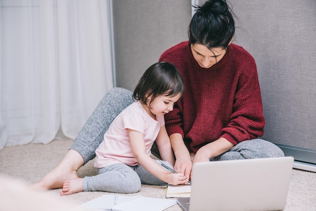 Moeder en haar kleine nieuwsgierige dochter werken thuis samen op een vloer en gebruiken een laptop en tekenen iets op het papier Zakenvrouw die in het huis werkt met kind