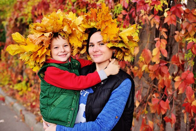 Foto moeder en jonge zoon dragen een kroon van gele bladeren knuffelen in de herfst park achtergrond glimlachen en kijken naar de camera.