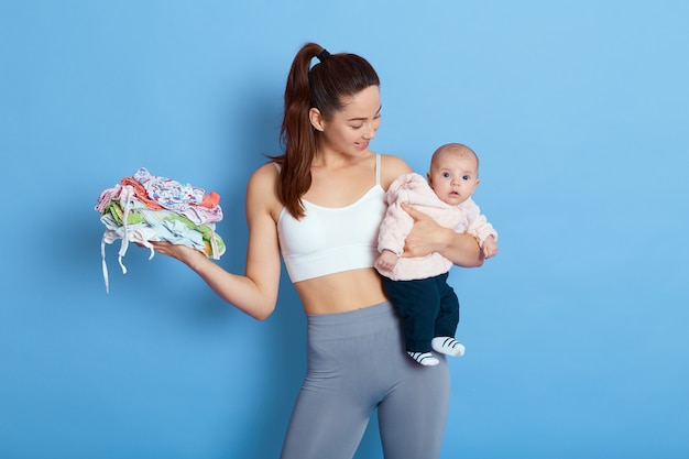 Moeder en kind baby jongen meisje binnen, mama kijkt naar haar pasgeboren dochter, houdt kinderkleding in de andere hand, geïsoleerd op blauwe achtergrond, aantrekkelijke vrouw met baby die sportkleding draagt.