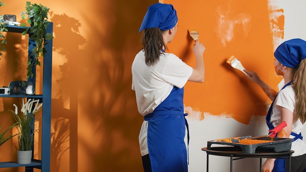 Moeder en kind gebruiken oranje verf om de kleur van de muren te veranderen en huishoudelijk werk te renoveren met doe-het-zelf gereedschap. kleine familie schilderij appartement ruimte met penseel en apparatuur, interieur.
