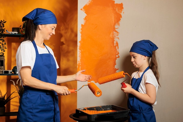 Moeder en kind schilderen huismuren met oranje verfkleur en rolpenseel, samen renoveren van appartementskamer. Mensen die plezier hebben met het opknappen van huishoudelijk werk, met behulp van diy-penseelgereedschap.