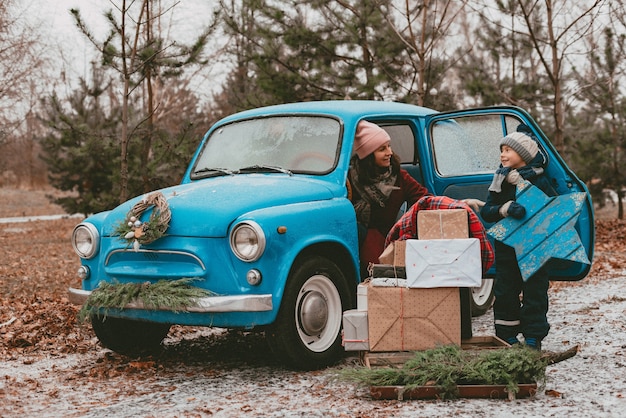 moeder en kind Versierd met blauwe retro auto met feestelijke kerstboomtakken, geschenkdozen ambachtelijk inpakpapier, een krans dennennaalden. Nieuwjaar familie-uitstapje. Kinderdroom, herinneringenverlangens.