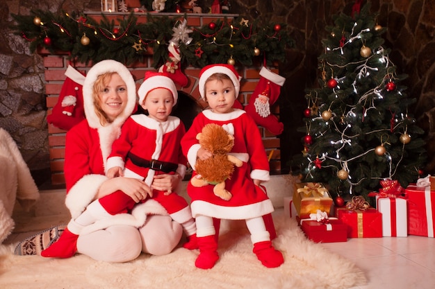 Moeder en kinderen zitten in de buurt van open haard en kerstboom met geschenkdozen. Broer en zus in kerstpakjes