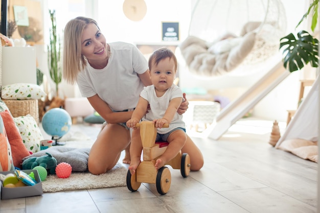 Foto moeder en kleine dochter brengen tijd door in de speelkamer klein meisje op een houten scooter moederlijke zorg liefde concept