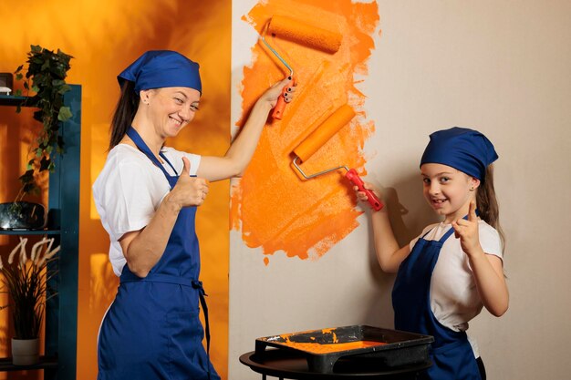 Moeder en meisje schilderen en geven duimen op, gebruiken oranje verf en rollerbursh om het appartement op te knappen. Mensen die het goed doen, houden van goedkeuringsgebaar en het renoveren van kamermuren.