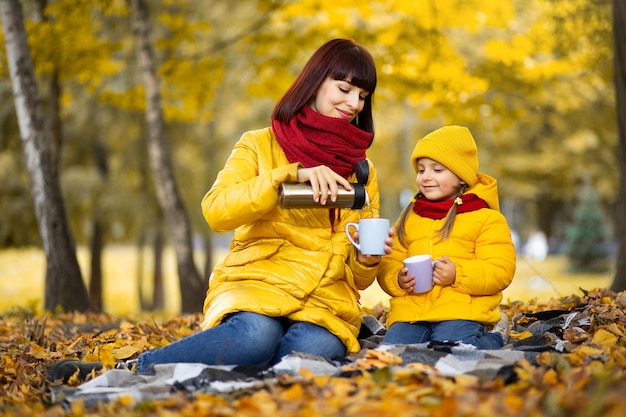 Moeder en schattige dochter, zittend op een deken in een prachtig gouden herfstpark. Mooie vrouw die hete thee uit een thermoskan in kopjes schenkt