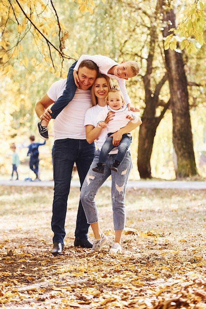 Moeder en vader houden kinderen op schouders en in handen. Vrolijk jong gezin maakt samen een wandeling in een herfstpark.